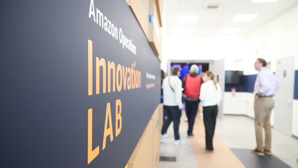A close shot of a wall of boxes that reads "Amazon Operations Innovation Lab", with a group of tour guests entering the facility.